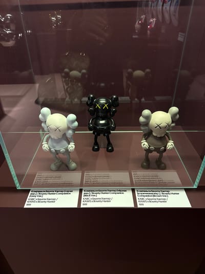 The image shows a display case containing three KAWS x Bounty Hunter Companion figures. The figures are in black, grey, and brown. Each figure is standing on a white label that has text printed on it in both Russian and English, identifying the item as "KAWS x Bounty Hunter" and the year "1999". The display case appears to be in a museum or gallery setting. The background is dark and out of focus, but shows reflections of other objects and possibly people. There is no information in the image to suggest that this is located in Moscow, Russia.