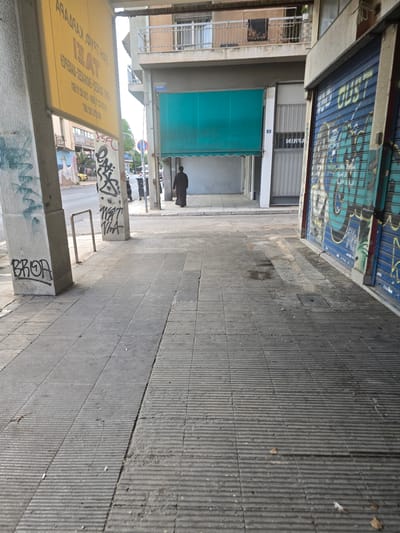 A lone figure, dressed in dark clothing and a hood, walks away from the camera on a paved sidewalk in Athens, Greece. The sidewalk is made of large, textured concrete slabs and shows signs of wear and some dampness. To the left of the figure, a prominent yellow sign with Greek lettering is attached to a concrete pillar. The pillar itself is covered in graffiti. Further to the left, another concrete pillar, also graffitied, supports a cantilevered walkway. In the background, a building with a blue, metal security gate covered in graffiti stands to the right. Adjacent to this is a storefront with a light-colored metal grate. Above, balconies with railings are visible on the building. The overall scene depicts an urban environment with elements of street art and everyday city life.