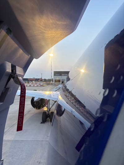 An airplane's open door reveals a red "REMOVE BEFORE FLIGHT" tag attached to a latch. The aircraft is parked on an airfield, with its engine and landing gear visible. In the background, airport buildings and lighting fixtures are illuminated, and the sky is overcast with hues of blue and orange, suggesting either dawn or dusk. The scene is set at Orio al Serio International Airport (BGY) in Orio al Serio, Italy.