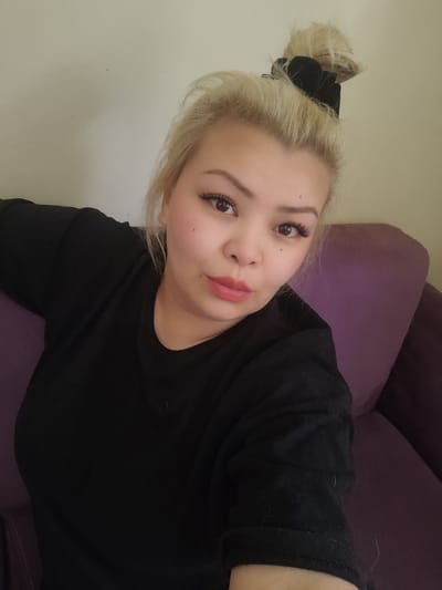 A woman with blonde hair pulled up in a messy bun with a black scrunchie is looking at the camera. She is wearing a black long-sleeved shirt and is sitting on a purple couch. She has on makeup, including false eyelashes and pink lipstick. The background is a light-colored wall. The scene is indoors, and appears to be a residential setting. No specific location in Sarıyer, Turkey is identifiable.