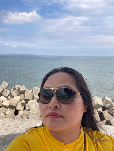 A woman wearing sunglasses and a yellow shirt takes a selfie with a body of water and a breakwater in the background. The breakwater is composed of concrete tetrapods. The sky is blue with scattered white clouds. The ocean appears calm. The location is the Davao City Coastal Road in Davao City, Philippines.