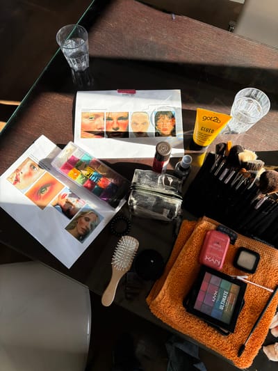 An overhead view captures a makeup artist's workspace on a dark, reflective tabletop, likely in an indoor setting within Paris, France, though no explicit Parisian markers are visible. Strong, directional sunlight illuminates the scene, casting distinct shadows.

Arranged on the table are numerous cosmetic items and reference materials. Two clear, faceted glass tumblers, each containing water, are present. Two white sheets of paper display printed images: one showcases four different makeup applications on faces with annotations, including a label "[make up]"; the other features close-ups of eyes and lips, alongside various model portraits.

Makeup products include a clear plastic case with 24 intensely colored square makeup pots, a yellow tube of "got2b glued" spiking glue, a small red-capped bottle, and a clear plastic spray bottle. A black fabric holder contains a dense collection of makeup brushes. An orange terry cloth towel serves as a base for an open pink compact of "NYX PROFESSIONAL MAKEUP Buttermelt Blush," a small square eyeshadow pan, and an "NYX ULTIMATE eyeshadow palette" displaying multiple colors. A white cotton swab and a black mascara wand rest on the towel.

Further objects include a clear, zippered, possibly worn, plastic makeup pouch, a wooden-handled hairbrush with white bristles, and a black spiral hair tie. No individuals are depicted within the frame, but the arrangement suggests preparation for a makeup application.