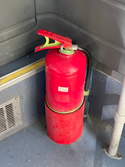 A red fire extinguisher with a yellow handle and green nozzle is mounted horizontally on a wall with a gray, speckled surface. The extinguisher is secured with a black hose-like attachment at the top. Below the extinguisher is a yellow and silver trim. The base of the wall features a metallic grate. Above the fire extinguisher is a handrail with its mounting hardware visible. The wall is gray and slightly curved. The setting may be within the confines of a public transport vehicle. The location cannot be ascertained as Yerevan, Armenia from the image.