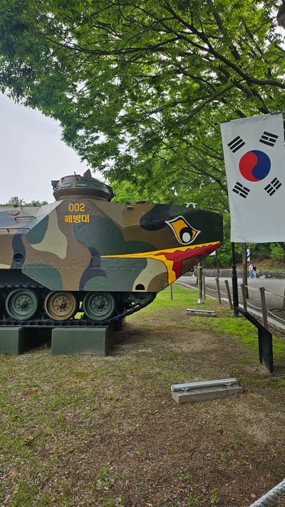 A military vehicle is displayed outdoors in Ulsan, South Korea. The vehicle is painted in a camouflage pattern of greens, browns, and black. The number "002" and Korean characters are painted on the vehicle's side, and a large eye-like graphic is painted on the rear. The South Korean flag is mounted behind the vehicle. The vehicle rests on green concrete blocks. The surrounding area is a park with grass and trees. A few people are walking in the background on a paved pathway.
