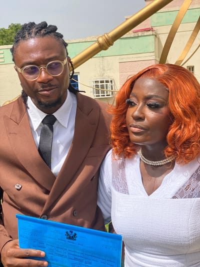 The image depicts a close-up, tilted shot of a couple, likely in Dadi Riba, Nigeria. The woman, in the foreground, has vibrant orange hair and is wearing a white lace-trimmed dress and a sparkling necklace. She has dramatic eye makeup and appears to be looking down, with a serene or thoughtful expression. The man is behind her and to the left, wearing a brown suit, white shirt, and a patterned tie. He has glasses with translucent frames and his hair is styled in dreadlocks. He is looking directly at the camera with a neutral expression. In his hands, he holds a bright blue document, which appears to be a marriage certificate. The text on the document is partially visible, indicating it's a "UNIFIED MARRIAGE CERTIFICATE" from the "FEDERAL REPUBLIC OF NIGERIA." The background shows a building with pastel-colored sections and windows, suggesting an outdoor setting with some architectural elements. The lighting indicates it is daytime.