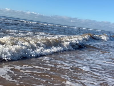 This image captures a coastal scene in Ciudad de la Costa, Uruguay, focusing on ocean waves under natural daylight. A prominent wave is breaking in the foreground, characterized by a crest of white foam and turbulent spray. The water immediately surrounding this breaking wave is a dark grey-brown, transitioning to a deeper blue-grey in the mid-ground where successive, smaller waves are visible receding towards the horizon. The immediate foreground consists of wet, dark brown sand partially covered by a thin layer of white, frothy water from receding waves. The sky, visible in the upper portion of the frame, is a clear, light blue with sparse white stratocumulus clouds concentrated near the distant horizon. No human subjects, animals, or artificial structures are present.