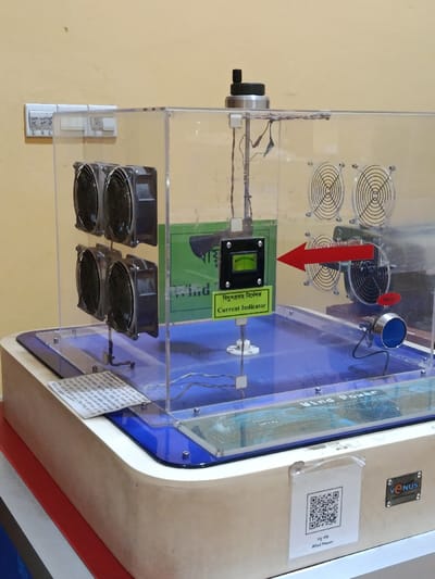 This image depicts a scientific demonstration exhibit, likely related to wind power, situated indoors against a plain light yellow wall in Dhaka, Bangladesh. The central subject is a clear acrylic enclosure housing various components.

Inside the enclosure, on the left side, four black computer-style fans are arranged in two vertical pairs, designed to generate airflow. On the right side, three smaller, silver-colored fans with protective grilles are visible, functioning as miniature wind turbines or generators. A prominent red arrow with "WIND DIRECTION" written in white points horizontally from the larger fans towards the smaller ones, indicating the intended airflow. Centrally positioned within the enclosure is a black square panel containing a green analog meter, labeled "Current Indicator" in both English and Bengali, mounted on a vertical rod. A green rectangular sign with Bengali text and "Wind Power" is partially visible behind the meter. Various wires connect the internal components.

The enclosure rests on a light-colored wooden or composite base with blue and white accents. On the front right exterior of the base, a white label with a black QR code and "Wind Power" in English and Bengali text is affixed. A small "VENUS" logo is visible on the far right of this label. On the front left exterior, a small white piece of paper with fine print, likely instructions or descriptive text in Bengali, is positioned under the acrylic lip.

In the background, mounted on the light yellow wall, is a white electrical switchboard containing multiple switches and outlets, with some switches marked "L" and "L L". No individuals are present in the scene. The overall setup suggests an educational or public display demonstrating the principles of wind energy generation.