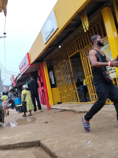 The image depicts a street scene in Umuode, Nigeria. Several people are present: a man in a black tank top and pants is walking on the right side of the frame, a woman in a patterned dress and another person in black are standing near a storefront on the left, and others are further down the street. The buildings are mostly yellow, with one storefront having a red awning. There is a sign that says "Connect Point" above one of the storefronts. The street surface appears to be paved but uneven, with a puddle of water visible. A motorcycle is parked along the street. A generator is near the puddle. A speaker is visible inside one of the stores.