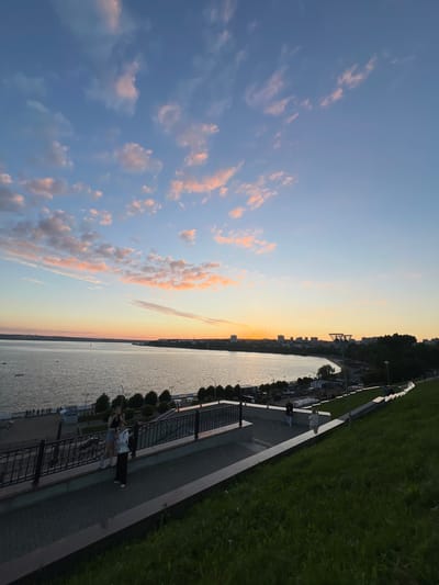 The image shows a scenic view of the Kama River in Izhevsk, Russia, at sunset. The sky is a vibrant gradient of blue, pink, and orange, with scattered clouds. The river is wide and calm, reflecting the colors of the sky. In the distance, the city skyline of Izhevsk is visible, with buildings and trees lining the shore. A paved walkway runs along the riverbank, with a black metal railing. Two young women are standing on the walkway, one taking a photo with her phone, and the other looking out at the river. The walkway is surrounded by a grassy slope, which is illuminated by the fading sunlight. There are streetlights visible along the walkway, and a few boats are dotted on the river. The overall atmosphere is peaceful and serene.