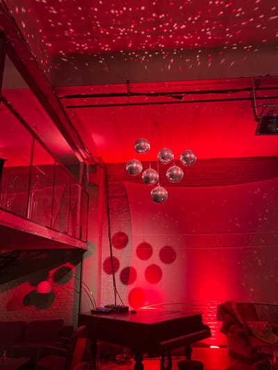 The image depicts an interior space within Roubaix, France, presenting as an event venue or lounge, illuminated predominantly by red ambient lighting. A metal-framed mezzanine with a transparent railing is situated along the left side of the scene. Walls feature exposed brickwork, partially covered by a large, light-colored fabric backdrop on the right. The ceiling exhibits an industrial design with visible structural beams and track lighting fixtures.

Seven mirrored disco balls are suspended at varying heights from the ceiling in the central-right area, reflecting the red light and projecting numerous small, shimmering red and white light points onto the ceiling and backdrop. An eighth, smaller disco ball is visible higher and further to the right. A dark-toned grand piano is positioned centrally in the foreground, with a small electronic mixer resting on its lid. Furniture includes a dark sofa partially visible on the far left and a plush red armchair with a draped blanket on the far right. A multi-headed floor lamp with a red shade is located beneath the mezzanine. Electrical cables are visible on the floor near the piano and running vertically along a wall support. No individuals are present in the scene.