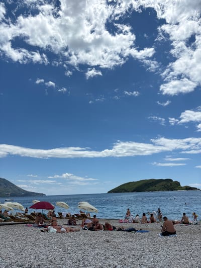 This is a wide-angle, full-frame, eye-level shot of a pebble beach in Budva, Montenegro. The sky is a vibrant blue with scattered, fluffy white clouds, some of which form a horizontal band across the mid-horizon. The sea is calm and blue, with a small, green, tree-covered island visible in the distance. The beach is populated with many people sunbathing, sitting, and relaxing on lounge chairs and towels. Several white umbrellas are set up for shade, along with one red umbrella. The people are mostly dressed in swimwear, and their activities include sunbathing, conversing, and looking at their phones. The pebble texture of the beach is prominent in the foreground. The overall impression is a lively and sunny beach scene on a summer day.