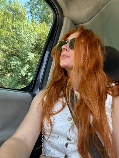 The media file shows a young woman with long, reddish-orange hair, wearing sunglasses and a white, lace-trimmed top. She is lying down inside a vehicle, her head propped up on the headrest. Her eyes are closed, and her lips are slightly parted, suggesting she is asleep or resting. The interior of the car, including the headrest and window frame, is visible. Outside the window, blurred green trees and a blue sky indicate that the vehicle is in motion, likely driving through a wooded area on a sunny day. The scene is oriented sideways, common for selfies taken with a phone held in a certain way. The woman's arm is also visible, with a tan line suggesting it has been exposed to the sun. The overall impression is one of a relaxed and peaceful moment during travel. The location is Medinski Krš, Montenegro. There is no visible text in the image.
