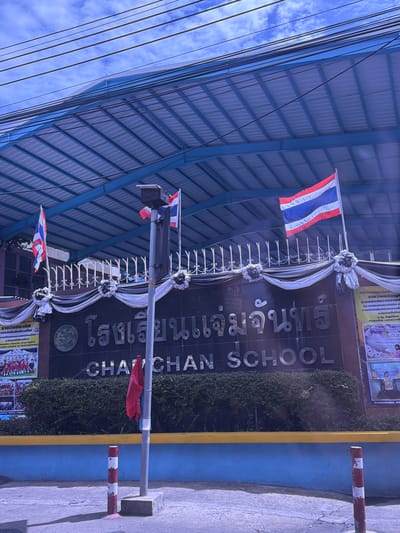 The image shows the exterior of "CHAI CHEN SCHOOL" in Bangkok, Thailand. The school building has a large, dark-colored facade with the school name prominently displayed in silver lettering. Above the school name, Thai lettering is also visible. The building is covered by a blue, corrugated metal roof supported by a metal frame. Three flags are visible: two Thai flags flying from poles on the roof, and a red flag attached to a lamppost in front of the school. The lamppost is gray with a solar panel on top. In front of the school is a manicured hedge. A yellow and blue painted wall runs along the bottom of the image, with a gray concrete sidewalk in front of it. Two red and white striped bollards are present on the sidewalk. The sky above is blue with scattered white clouds, and overhead power lines are visible. There are posters on the right side of the building, with text and images that are partially visible. One poster appears to depict students in uniforms. The image has a bright, sunny feel, though the lighting suggests it may be mid-day.