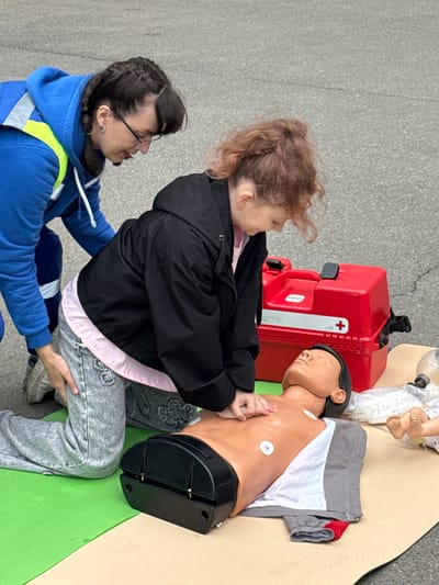 The image appears to depict a first aid training exercise or demonstration. A young person with auburn hair is positioned over a mannequin, with their hands placed on the chest in a posture suggestive of performing chest compressions. Another individual, wearing a blue hooded sweatshirt with a yellow reflective stripe, is assisting and holding the young person's leg or torso. The mannequin lies on a green and beige mat. Behind the young person, a red first aid kit or medical case is visible. The setting appears to be an outdoor space, potentially a paved parking lot or similar area. There is nothing in the image that specifically locates it in the city of Dmitrov, Russia.