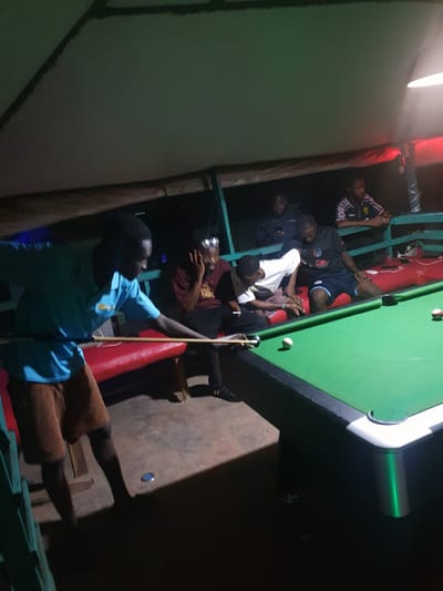 In Lokoja, Nigeria, a group of young men are gathered around a pool table in an outdoor, dimly lit recreational area, possibly an open-air bar or lounge. The scene is illuminated by a mixture of overhead lighting, casting shadows and highlighting the green felt of the pool table.
The focal point is a man in a turquoise polo shirt and brown shorts, actively playing pool. He is positioned to the left, leaning over the table with a pool cue extended, preparing to strike a white ball. His gaze is concentrated on the game.
Other individuals are dispersed around the table, observing the game or engaged in conversation. To the right of the player, a man in a maroon t-shirt and black shorts is seated, with his hand to his face as if in thought or amusement. Further back, several other men are visible. One, in a black t-shirt with a red emblem and blue shorts, is seated, his attention directed towards the pool table. Another man, wearing a black and white patterned sports jersey, is also seated, looking towards the game. Two other individuals are partially visible in the background, one appearing to be looking at his phone.
The environment suggests a casual, social gathering. A red bench runs along the side of the pool table, and the flooring appears to be concrete or tiled. The overhead structure, possibly a thatched roof or canopy, is visible, contributing to the outdoor setting. The overall atmosphere is one of relaxed camaraderie, centered around the game of pool.