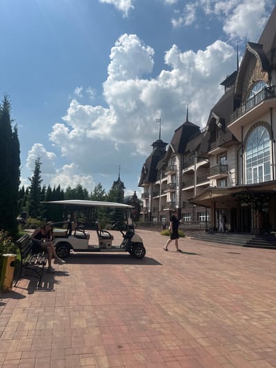 In Bogdanikha, Russia, a resort complex is captured on a sunny day with a bright blue sky filled with cumulus clouds. In the foreground, a beige golf cart is parked on a red brick pathway. To the left of the golf cart, a young woman with blonde hair wearing a black tank top and shorts sits on a park bench, looking down at her phone. To the right of the golf cart, a person in a black dress and sandals walks away from the camera towards a large, ornate building with multiple balconies, arched windows, and dark, sloping roofs. The building appears to be a hotel or resort. In the background, numerous tall evergreen trees surround the area, and a dark, spire-like structure is visible behind the main building. The overall scene depicts a serene and luxurious outdoor environment.