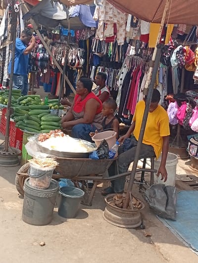 The image captures a vibrant and busy outdoor market scene during daylight hours, likely under a sunny sky given the bright illumination and shadows. Several individuals are visible amidst various stalls, some selling food and others clothing.
In the foreground and midground on the left, a stall displays a large quantity of fresh green cucumbers neatly stacked. Behind this stall, a man in a blue t-shirt and light-colored pants stands, appearing to be looking upwards or adjusting something with his hands near his head, possibly engaging with a phone or an overhead structure. To his right, two women and a small child are seated on low stools. The woman in the foreground wears a red top, while the other woman wears a lighter-colored top. Both are looking towards the right side of the frame. The child, dressed in a black tank top, sits in front of the woman in red, looking down or with eyes closed.
Central to the midground is a modified wheelbarrow acting as a vendor's stand. It holds a large, round metal basin filled with a light-colored, possibly rice-based, cooked food, topped with some darker ingredients. A plastic bottle of Aquafina water rests on the wheelbarrow. Nearby, on the ground, are several buckets: a large grey bucket, a smaller blue bucket filled with water, and a clear plastic bucket containing numerous skewers of what appear to be small dried fish or snacks.
To the right of the wheelbarrow, a young man in a yellow t-shirt and dark pants is seated on a stool, leaning against a wooden pole that extends upwards, likely supporting an umbrella or awning. At his feet, an old metal wheel rim is repurposed to hold several whole coconuts or similar produce. Black plastic bags are also visible on the ground near him.
In the background, behind the various vendors, numerous clothing racks are packed with garments of different styles and colors, including dresses, shirts, and what appear to be bras on hangers. Some children's clothing with cartoon patterns (like teddy bears) can also be seen hanging. Large, brown-colored awnings or market umbrellas provide shade over the stalls. Faint text of "Coca-Cola" is visible on a red cooler or stand in the far left background. The overall atmosphere suggests a bustling commercial environment.