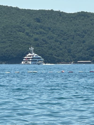 In Boreti, Montenegro, a large white yacht with multiple decks is sailing on calm blue water, creating a visible wake. Behind the yacht, a dense, green, tree-covered hill slopes down towards the water. In the foreground, several buoys are spaced out across the water. A structure resembling a building with a long, flat roof is visible on the shoreline in the distance, partially obscured by the yacht. The sky is a pale, clear blue.