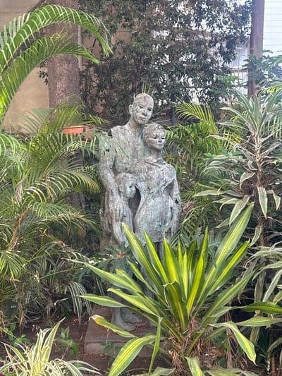 This image shows a bronze statue of two figures, one taller than the other, embracing and partially covered by foliage. The statue is located in a garden setting with lush green plants surrounding it. The taller figure appears to be an adult, with the smaller figure possibly representing a child or a younger person. The statue has a weathered appearance, typical of bronze exposed to the elements. The background includes trees and a building with windows, suggesting an urban or suburban environment. Based on its location, this statue is likely situated in the Garden of the Fallen Soldiers in Ramat Gan, Israel, a memorial site dedicated to soldiers who died in service.