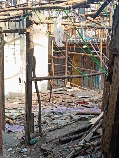 A devastated interior space features exposed wooden beams and supports, suggesting collapse or demolition. Debris litters the ground, including wood scraps, torn fabric, and unidentified refuse. A white, draped object hangs from the overhead structure. A glimpse of a distant, makeshift building shows corrugated metal and rough wood construction. The overall impression is one of destruction, poverty, and makeshift repair.