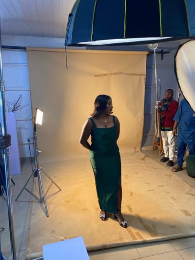 In Bukuru, Nigeria, a woman in a form-fitting, dark green, floor-length dress with a thigh-high slit stands posing in a studio setting. She wears black open-toed heels with embellished straps. Her hands are clasped behind her back, and she looks to her right, away from the camera. The studio has a light beige backdrop that appears to be paper or fabric, which is taped to the floor in several places. Several studio lighting equipment pieces are visible, including a large softbox light and a smaller LED panel light on a stand. Two men are present in the background; one stands to the right of the woman, wearing a red jacket with green and blue stripes, white pants, and black shoes. Another man, partially obscured, stands further to the right in a blue garment. A metal light stand is in the foreground to the left, and a white board is on the floor in the bottom right corner. The overall lighting is bright, characteristic of a professional photo shoot.