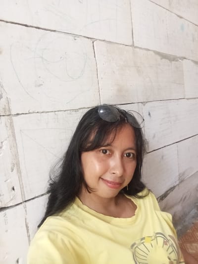 A woman wearing a yellow shirt and sunglasses is posing for a selfie. She is positioned against a wall constructed of what appears to be white bricks or blocks. Faint drawings or markings are visible on the wall. The location is possibly Baron, Indonesia.