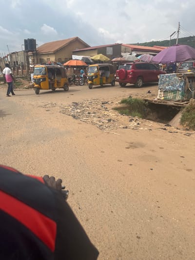 A medium shot captures a street scene in Abuja, Nigeria, on a cloudy day. The foreground is dominated by a portion of a person's arm, clad in a black and red jacket, holding onto handlebars that are not visible. The road surface is unpaved and appears dusty, with scattered debris and some puddles or stains.
In the mid-ground, a yellow auto-rickshaw, known locally as a 'Keke Napep', is parked on the right side of the road, facing left. Another similar auto-rickshaw is visible behind it, also yellow. To the left, a person wearing a white shirt with a pink accent and dark trousers walks away from the camera. Behind the auto-rickshaws, a red SUV is parked. Several market stalls are visible in the background, with various colorful umbrellas providing shade. These stalls are constructed from corrugated metal and wood. A large purple umbrella is prominently displayed over one of the stalls. In the far background, a hillside covered in vegetation rises under the overcast sky. A large water tank on a metal structure is situated to the left of the buildings. The overall environment suggests a bustling, informal market area.
