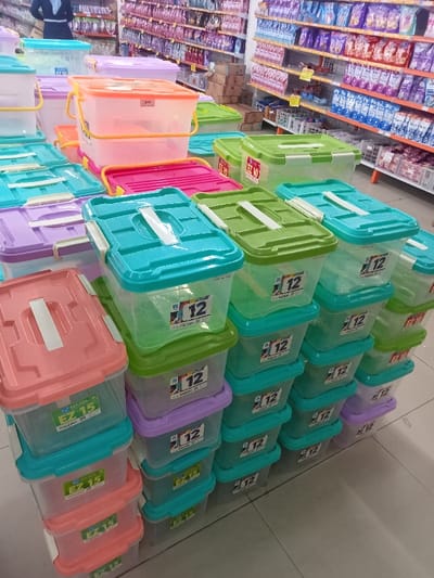 The image shows a display of plastic storage containers stacked on top of each other in a store in Pati, Indonesia, located at latitude -6.757 and longitude 111.039. The containers are various sizes and colors, including blue, green, pink, and lavender. The containers are labeled with the words "EZ 12" and "EZ 15", suggesting that they are for storing items. There are several containers visible in the image. The containers are arranged in a neat and orderly fashion. The store floor is tiled, and the background includes a wall of shelves stocked with various items.  There are no people visible in the image.  There is a small, rectangular, white price tag near the top of the stack.