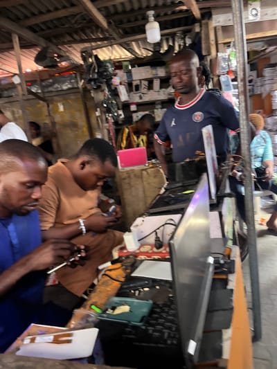 This image depicts an active electronics repair and sales stall within a bustling market in Lagos, Nigeria, characteristic of areas like Computer Village in Ikeja. The overhead structure consists of exposed wooden beams and corrugated metal roofing, illuminated by a single white LED bulb.

Four individuals are primarily visible. In the left foreground, a dark-skinned male in a blue garment is intently focused on repairing a smartphone with a small tool. To his immediate right, another dark-skinned male in a brown top, wearing a white wristband, is similarly engaged in examining or repairing a separate smartphone. Both appear actively involved in detailed work.

In the mid-ground, a bald, dark-skinned male wearing a blue Paris Saint-Germain (PSG) jersey stands, hands obscured, observing the scene with a downward gaze. Further in the background, a person in a yellow and black patterned garment is partially visible, leaning over a surface, and a female in a light blue top is partially visible to the far right.

The workstations are densely cluttered with electronic components, wires, tools, a white power adapter, and a red multi-socket extension cord. A disassembled laptop screen is prominently positioned vertically in the mid-right foreground. Shelves lining the rear wall are stocked with various electronic accessories, phone cases, and other merchandise, indicating both repair services and retail sales. The overall environment is functional, utilitarian, and indicative of high commercial activity.