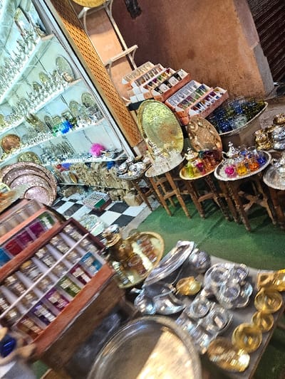 The image depicts a richly stocked market stall, likely within a souk in Marrakesh, Morocco. The scene is dominated by an extensive display of traditional Moroccan decorative items, primarily metalware and glassware.
On the left, a tall, multi-shelf glass display case is filled with numerous silver-colored metal plates, trays, and possibly teapots or pitchers, meticulously arranged. Below this case, additional metal trays and small items are visible, alongside a small, fluffy pink decorative object. Further into the background on the left, a black-and-white checkered floor pattern is discernible.
Towards the foreground on the left, a wooden display unit with multiple compartmentalized sections showcases rows of small, colorful items, possibly bottles of perfumes, oils, or spices.
On the right side of the image, various goods are presented on low wooden tables or stools and directly on what appears to be a green-carpeted floor area. These items include: large, ornate brass or gold-colored trays; smaller silver-colored trays holding sets of vibrantly colored tea glasses (red, green, blue, yellow, purple) accompanied by silver-colored teapots; and an oval-shaped, textured decorative object. Stacked cardboard boxes containing rows of clear and colorful small glasses are also prominently displayed on the right, against a reddish-brown wall.
The foreground on the right features a collection of silver-colored and gold-colored metal objects, including trays, bowls, and possibly components of tea sets, spread across a surface.
No individuals are visible in the frame. The focus is entirely on the merchandise and its arrangement within the retail environment.