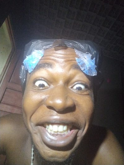 A close-up, low-light, upward-angled selfie of a Black male is shown. The man has dark skin and is wearing a clear plastic shower cap on his head, with two blue, ice-pack-like objects attached to the top of the cap on either side of his forehead. He has wide, staring eyes and a large, open-mouthed smile, revealing his teeth. His neck and upper chest are visible, and he appears to be wearing a necklace of light-colored beads. The background is dark and appears to be indoors, with some textured tiling visible on the ceiling. To the left of the man, a portion of a wooden door or frame is visible. The image appears to have been taken in or around Ihiagwa, Nigeria.