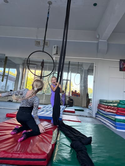 In a brightly lit indoor space, possibly a gymnasium or dance studio, two young girls are engaged in aerial acrobatics. The setting features high ceilings, large windows that allow natural light to filter in, and what appears to be a mirrored wall reflecting the scene. The floor is covered with padded mats in red, green, and blue, suggesting a safe environment for athletic activities. Several gymnastic apparatus are visible, including large hoops and black aerial silks.
One girl, with blonde hair styled in a braid adorned with a white flower, is dressed in a leopard-print top and black leggings. She is leaning on a red mat, her body positioned as if mid-performance or practicing a pose. Her facial expression is not clearly visible.
The other girl, who appears younger, is suspended mid-air by black aerial silks. She is wearing a vibrant purple leotard and has an excited, open-mouthed expression, suggesting joy and enthusiasm. Her blonde hair is loose, and she is gripping the silks with her hands.
The overall atmosphere seems energetic and playful, characteristic of children practicing a sport or performance art. The presence of gymnastic equipment and the girls' attire indicate a training or practice session. The time of day appears to be daytime, as suggested by the natural light coming through the windows. The location is identified as Dnipro, Ukraine. No visible text is present in the image.