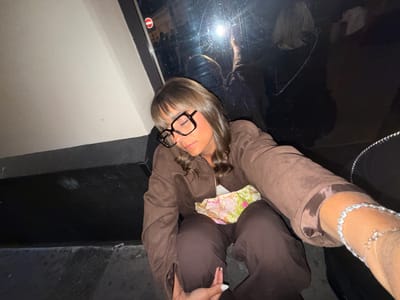 A woman with shoulder-length blonde-brown hair is sitting on the ground, possibly on a sidewalk, with her eyes closed. She is wearing large, dark-framed glasses, a brown jacket, and matching brown pants. A floral-patterned garment or bag is visible between the jacket and pants. She is holding her hands near her lap, and a bracelet is visible on her outstretched arm.
Behind her is a reflective window with a no-entry sign in the upper left corner. Reflections of people can be seen in the window. The ground appears to be asphalt or concrete.
Without additional context, it is difficult to confirm whether the image was taken specifically in Nice, France.