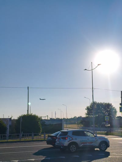 The image depicts a bright, sunny day in the city of Sirius, Russia. The sky is a clear, pale blue, with the sun a prominent, overexposed white disc in the upper right quadrant, creating lens flare effects. An airplane is visible in silhouette, flying in the mid-ground against the blue sky.
In the foreground, a paved road with white lane markings is visible. Two cars are parked or driving on the road. The car on the right is a silver SUV with the text "АВТОШКОЛА МАСТЕР" (Driving School Master) and a phone number "295-70-70" printed on its side. It also features a red and blue checkered logo. The car to its left is a dark-colored sedan.
Behind the cars, a white metal fence runs horizontally across the mid-ground. Beyond the fence is a strip of green grass and then a line of trees, the largest of which is a full, green deciduous tree in the mid-right of the frame. In the background, there are various structures and utility poles, including a tall street lamp with two lights on curved arms. A pedestrian crossing sign with a yellow background and a black pedestrian symbol is visible near the right edge of the image. The overall scene suggests an urban or semi-urban environment during daylight hours.