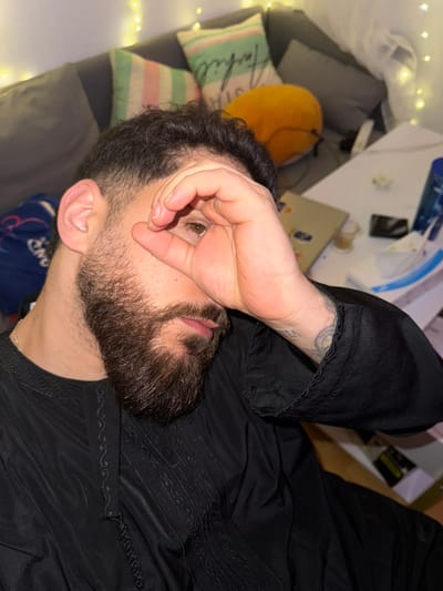 A man is depicted in a close-up, high-angle shot, lying down indoors, possibly on a couch or bed. He is positioned with his head towards the bottom of the frame and his body extending upwards to the right. His right hand is raised, partially covering his face, with his thumb and index finger forming a circle, allowing his right eye to peek through. His visible eye is brown. He has dark, curly hair, a full beard, and a mustache.

He is wearing a dark, likely black, long-sleeved garment that features intricate, geometric embroidered patterns on the chest and lower sleeve areas. A tattoo with a circular design and faint text or numbers is visible on his left forearm, just beneath the cuff of his sleeve.

The background suggests an indoor setting, likely a casual living space or bedroom. To the left of the man, several cushions are visible. One is striped in light green, pink, and white, with partially visible text that appears to spell out "Farfil" or similar. Another cushion is a solid orange or yellow color. Behind and above the man's head, a light-colored surface, possibly a table or desk, holds various items including papers and what looks like a mobile phone. String lights, often referred to as fairy lights, are draped in the top-left and bottom-left portions of the frame, emitting a warm, soft glow, suggesting the photo was taken during the evening or at night. The overall impression is one of a relaxed, intimate moment.