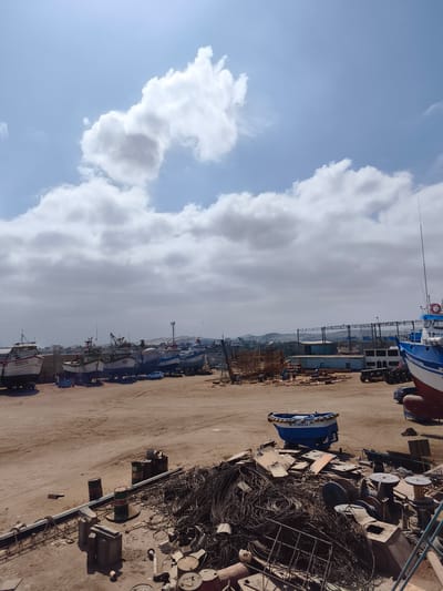The image depicts a ship repair yard in Chimbote, Peru, under a cloudy sky. In the foreground, the ground is covered with various industrial debris, including a large pile of tangled wires, metal plates, barrels, and spools of wire. Several boats are visible in various states of repair or dry-docking in the mid-ground, with some resting on stands. A small blue and white boat, labeled "MI AURELIA," is prominently positioned in the middle. In the background, more ships and port structures are visible, including cranes and buildings. The overall scene suggests active, albeit somewhat unorganized, maritime industrial activity.