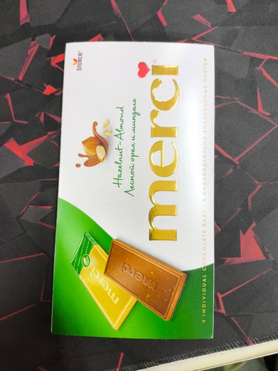 A box of Merci chocolates lies on a black and red patterned surface. The box is predominantly white with green accents and features a large, gold, embossed "merci" logo. Text on the box indicates "Hazelnut-Almond" and in Russian "Лесной орех и миндаль" which translates to "Hazelnut and almond". Below the main logo, smaller text reads "4 INDIVIDUAL CHOCOLATE BARS". Two individual chocolate bars are partially visible at the bottom of the box, one wrapped in yellow foil with "merci" printed in white, and another unwrapped chocolate bar with "merci" embossed on its surface. The scene is an indoor setting, and there are no people present. The location is not specified in the image.