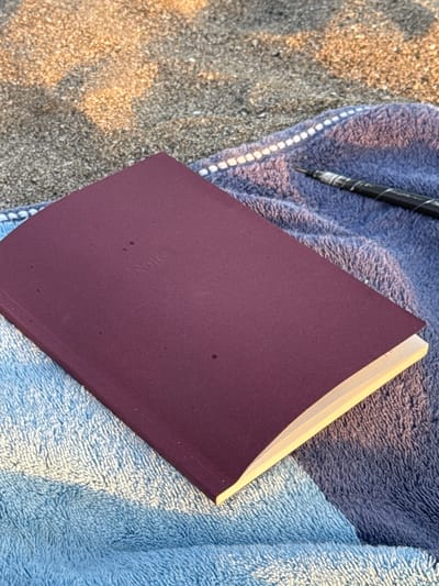 A closed, dark red notebook with the embossed word "Notes" on its cover lies on top of a blue towel. To the right of the notebook, a black and white marbled pen rests on the towel. The towel is partially on a sandy surface, illuminated by soft, warm light. The image is taken from a high angle, looking down. The location appears to be a beach in North Kuta, Indonesia, given the sand and towels indicative of a beach setting. There are no people visible in the frame.