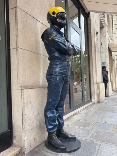 The image shows a life-sized statue of a person in a dark blue pilot's suit and a yellow helmet with a visor, standing with arms crossed in front of a building with large glass windows. The statue is positioned on a circular black base. The building appears to be a retail store, with "BREITLING" visible on the glass windows in gold lettering. To the right of the statue, a person wearing a black puffer jacket and a black baseball cap is standing, appearing to be on a phone call. The scene is on a paved sidewalk with large stone tiles. The architecture of the building suggests it is located in a European city, and the presence of the Breitling store indicates a commercial area. Based on the architecture and the Breitling store, the location is Paris, France, specifically on Rue de la Paix. The lighting indicates it is daytime.