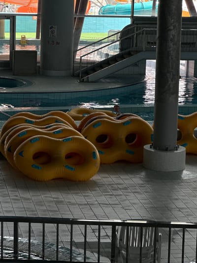 An indoor water park in Minsk, Belarus, features a swimming pool with inflatable tubes stacked on the tiled floor. A person is visible in the pool. In the background, a staircase leads to a waterslide. A pillar with a depth marker indicating "1.3 M" stands near the pool.