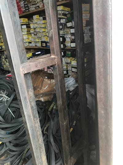 The image depicts the interior of what appears to be an automotive parts storage area or shop in Akure, Nigeria. The scene is viewed through the dark, rusted metal frame of a ladder or shelving unit positioned in the immediate foreground, obscuring parts of the view. To the left and bottom-center, large quantities of black automotive V-belts, many with yellow identification markings, are loosely piled on the floor. Some white fibrous material, possibly rope, is intertwined with the belts on the far left. Directly behind and above the piled belts, shelves are densely packed with numerous small cardboard boxes, primarily yellow, beige, and white, stacked in rows and columns. Visible labels on some boxes include "SKF" and "CFB," indicating automotive components like bearings or brake pads. Additional brown cardboard boxes are visible at the lower-center. The right side of the frame shows a plain white vertical surface. No individuals or specific actions are visible within the frame. The area appears to be a busy storage space for various vehicle components.