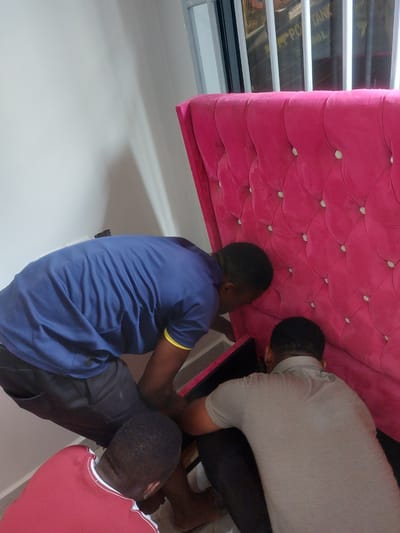 The image displays an indoor scene in Dar es-Salaam, Tanzania, where three adult males are focused on a large piece of pink upholstered furniture. The furniture, a tall item featuring deep button tufting with light-colored buttons, is positioned upright against a plain, light-colored wall.
On the left, a male wearing a navy blue short-sleeved t-shirt with yellow trim on the sleeve and dark trousers is bent over, facing downwards towards the base of the pink furniture. To his right, a second male, clad in a grey short-sleeved collared shirt and dark trousers, is also bent over, concentrating on the lower section of the furniture. Partially visible in the bottom left, a third male wearing a red short-sleeved t-shirt with white trim is likewise bent over, positioned near the feet of the individual in navy blue. All three individuals appear to be engaged in an activity at the base or rear of the furniture, possibly assembly or adjustment.
In the upper right background, a window is visible, secured by vertical white security bars. Through this window, a dark object with white text can be partially seen, displaying characters consistent with "PO TANK" and "OOL," indicating an external industrial or storage context. The immediate environment consists of light-colored walls and a light-colored floor.