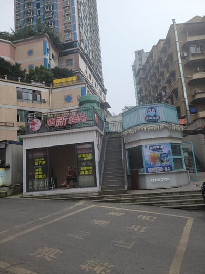 The image captures a street scene in Shapingba District, China. A building with a blue and pink facade is visible, along with a business marked by a red logo with Chinese characters and another building with a green air vent on its roof. Adjacent to the road is a small shop with white walls and large windows displaying Chinese text, with a person visible inside. Further down the street, a staircase leads up to a blue-and-white establishment labeled "QI.E ICE," featuring a penguin logo and promotional materials. The street surface is asphalt, with faded yellow painted markings and Chinese characters. The overall atmosphere is somewhat hazy.
