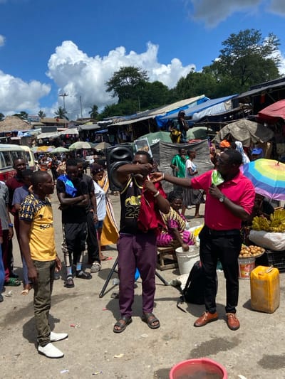 The scene is a bustling market area in Dar es Salaam, Tanzania, located at the coordinates -6.787 latitude and 39.113 longitude. A group of men and women are gathered around a large speaker, possibly selling merchandise or engaging in a performance. The man in the pink shirt is standing in front of the speaker, addressing the crowd. He is holding a microphone, and several people are looking at him attentively. The man wearing a purple shirt and a basketball jersey is holding a red cloth in his right hand. Several other individuals are standing nearby, observing the activity. Behind the group, there are stalls and vendors selling various goods. The market stalls have a variety of colorful fabrics, clothing, and goods. The overall atmosphere of the scene is bustling and vibrant, a typical sight in a Tanzanian market.