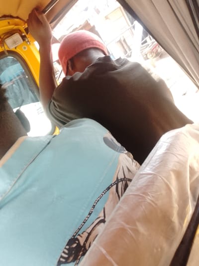 The image presents an interior view of a yellow motorized tricycle, located in Sabon Tasha, Nigeria. In the immediate foreground, the partial back and shoulder of an individual wearing a light blue shirt with a dark patterned design are visible on the left. To the right, a leg or object is partially covered by a translucent white plastic material. Positioned in the middle ground, another individual is seated facing away from the camera, wearing a dark short-sleeved shirt and a red knitted cap. This person's right arm is raised, with the hand gripping an overhead metal bar integrated into the tricycle's yellow interior panel. A small, rectangular white light fixture is mounted on the yellow ceiling panel above the seated individual. The background, viewed through the open side of the vehicle, is brightly lit and out of focus, depicting an urban outdoor setting during daylight.