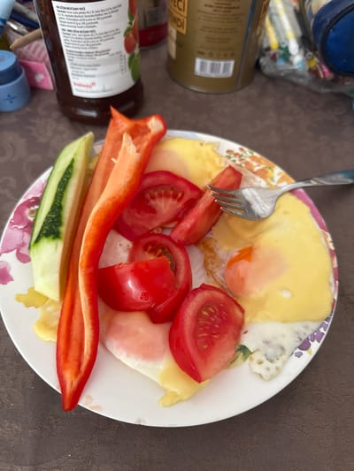 In Budva, Montenegro, a plate of food sits on a brown tablecloth. The plate contains fried eggs, sliced tomatoes, cucumber sticks, and red bell pepper strips. A fork rests on the plate, partially covering one of the fried eggs. In the background, several bottles and containers are visible, including a bottle with a label featuring fruit. The scene appears to be an indoor setting, possibly a dining area.