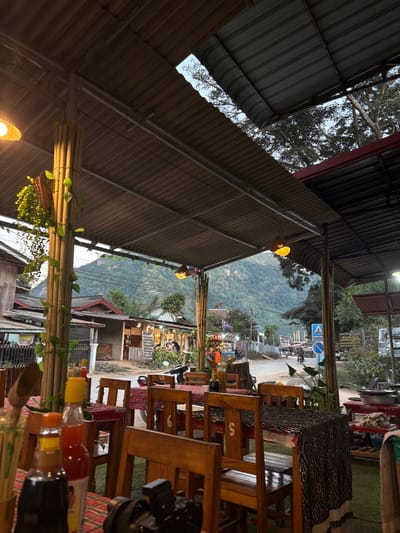 The image depicts a semi-open-air restaurant or eatery patio in Nongkhiao-Nua, Laos, at what appears to be late afternoon or early evening. The scene is viewed from a low angle within the establishment.
The foreground features multiple wooden tables with matching wooden chairs. Several tables are covered with red and white checkered tablecloths, while others have dark, patterned fabric. On the closest table to the viewer, a black camera body with a lens is partially visible, alongside three condiment bottles: one with a red cap, one with an orange cap, and a dark brown bottle. A cylindrical holder containing chopsticks or skewers is also present.
The restaurant structure consists of a corrugated metal roof supported by steel beams and columns clad in bamboo poles. Green vining plants are wrapped around these bamboo columns. Yellow, basket-style pendant lights are illuminated and hang from the roof structure. Gaps in the roof system reveal a bright sky and the canopy of trees overhead.
Beyond the immediate restaurant seating, a paved street runs through the town. Buildings, primarily single-story structures with corrugated metal roofs, line both sides of the road. Lighting is visible within some of these buildings. A white scooter is parked on the left side of the street. Several indistinct human figures are visible on the street; one is near the scooter, and others are further in the distance. On the right side of the road, a blue rectangular road sign with a white arrow pointing left is visible, accompanied by a smaller white rectangular sign below it.
In the far background, a range of large, green, tree-covered mountains rises prominently under a bright, possibly overcast, sky. The overall scene suggests a quiet, rural town setting.