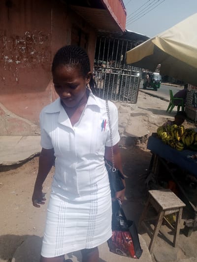 The image depicts an outdoor street scene in Aba, Nigeria, under bright daylight. A young adult female, of African descent, occupies the foreground, positioned slightly left of center. She wears a white, short-sleeved collared uniform comprising a pleated top and a matching knee-length skirt. Two pens, one blue and one red, are clipped to her left shirt pocket. Her dark hair is styled in braids, pulled back. She carries a black shoulder bag with a silver chain strap over her left shoulder and a black and red patterned shopping bag in her left hand, which is held close to her body. Her head is tilted downwards, and her gaze is directed towards the ground.
The ground beneath her is unpaved and appears to be dusty earth or dirt. To the immediate left and behind the woman, a reddish-brown building wall is visible, exhibiting some patches of lighter-colored surface. A dark metal grate or fence structure is situated further back on the left side.
To the right of the woman, a beige or off-white umbrella provides shade over a makeshift market stall. The stall consists of a wooden structure topped with a blue cloth, upon which a pile of ripe yellow bananas is displayed. A person, largely obscured and seated on a low stool or ground behind the stall, is visible. A small, rustic wooden stool is positioned on the ground in front of the stall. In the background, a paved road is visible, with a green three-wheeled vehicle (auto-rickshaw type) driving on it. Another individual is partially visible sitting on a green plastic chair on the roadside further back.