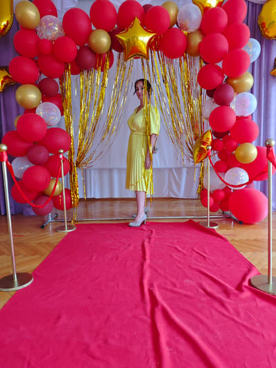 A woman in a yellow dress stands on a red carpet in front of a balloon arch. The arch is decorated with red, gold, and white balloons, as well as gold tinsel. Two stanchions with red rope borders the carpet. The woman is wearing gray shoes. The location is a room in Markovskii, Russia with a wooden floor and white curtains.