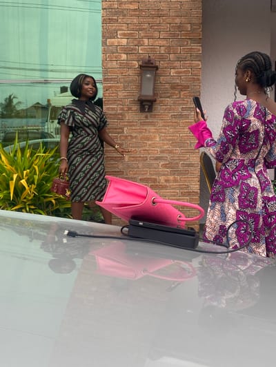Two Black women are standing outside a building with a brick facade and large glass windows. The woman on the left is wearing a patterned dress and holding a small red handbag. She is looking towards the right, away from the camera. The woman on the right is wearing a colorful patterned dress and holding a smartphone in her right hand, appearing to take a photo or video. She is facing away from the camera, towards the woman on the left. In front of the woman on the right, on the ground, is a large pink bag. A grey car is visible in the foreground, with its reflection on the surface. The environment suggests an urban setting, potentially Ado, Nigeria, given the architectural style and vegetation.